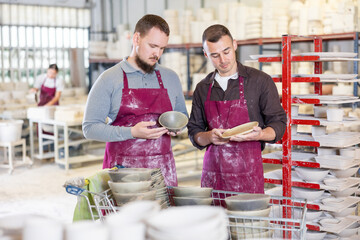 Two enthusiastic young male potters in stained burgundy aprons examining handcrafted ceramic wares in studio and exchanging ideas about techniques of casting
