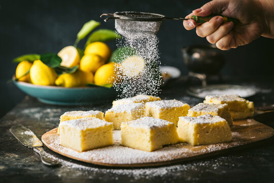 Side angle action shot of female hand sprinkling powdered sugar over lemon cake squares, bowl of lemons in BG