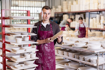 Young man in uniform posing with blanks of ceramic products in factory
