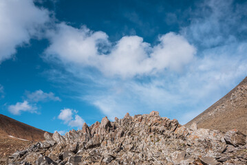 Sunlit stone hill in high mountain valley under clouds in blue sky. Pile of stones on mountain pass in bright sun. Rocky hill of round shape in sunny day. Stony mountain top in changeable weather.