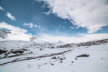 Cold snow-white landscape with snow-covered pointy peak under clouds in blue sky. Snowy rocky...