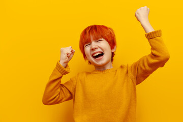 happy red-haired teenage boy celebrating victory on yellow isolated background, child winner with...