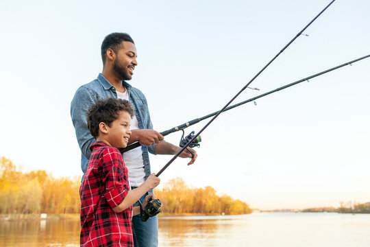African American man and child sitting on a wooden pier holding fishing rods on the river, dad teaching his son to fish in the lake, family resting and relaxing on the weekend