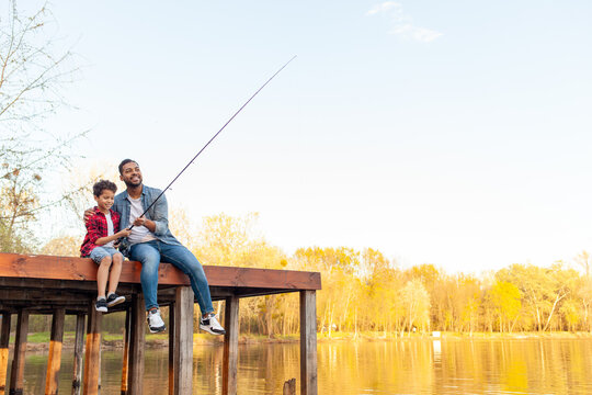 African American man and child sitting on a wooden pier holding fishing rods on the river, dad teaching his son to fish in the lake, family resting and relaxing on the weekend