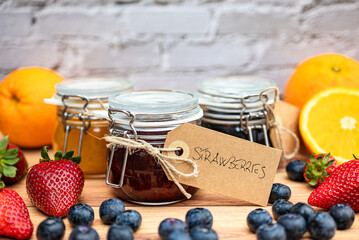 Glass jars with homemade jam decorated with handmade brown cardboard labels attached with string. In the foreground, a jar of homemade strawberry jam, in the background, blueberry and orange jam.