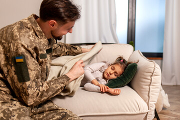 happy soldier of Ukrainian army in camouflage uniform returned home and puts his daughter to bed,...