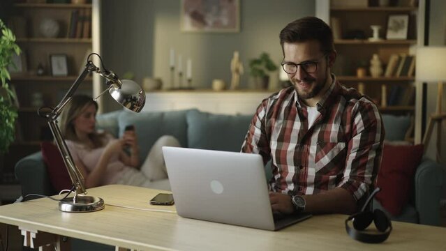 Happy smiling freelancer working from home on the laptop while Sitting on the desk in stylish cozy living room. Woman using mobile phone in the background