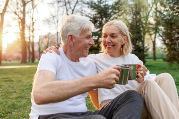 elderly couple of seniors man and woman hold metal cups and drink tea in the park outdoors, gray-haired grandparents communicate and hold mugs in nature