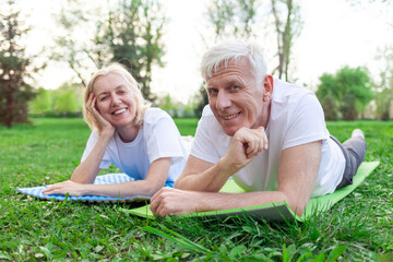 elderly athletic couple seniors man and woman lying on yoga mat and relaxing in park outdoors, gray-haired grandparents doing yoga and meditating and smiling in nature
