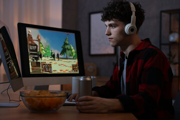 Young man with energy drink and headphones playing video game at wooden desk indoors