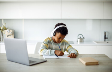 Seven year old mixed race boy doing homework with laptop and headphones.