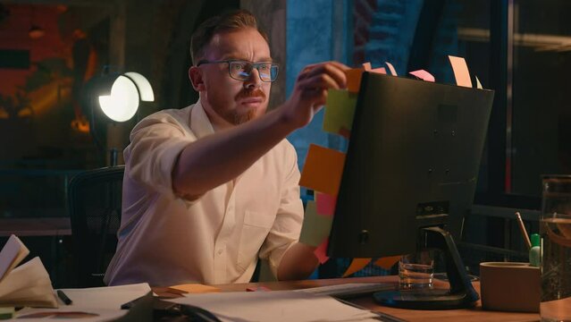 Caucasian man business guy businessman male in dark night evening office work reading check information on stickers throw away dissatisfied read sticky paper notes on computer monitor screen deadline