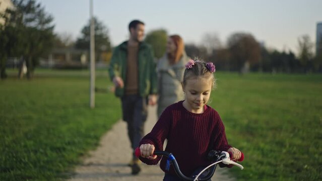 Little girl riding bike outdoors in city park with parents walking watching in background