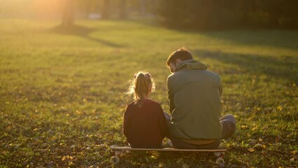 Back view of young dad and little daughter sitting on the skateboard while girl is playing with leaves in the autumn park - Powered by Adobe
