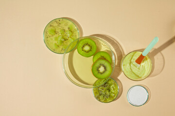 A glass cup with yellow body scrubs and a plastic stick inserted, next to a glass tray filled green essence and kiwi slices, a dish of chopped kiwi and the other with white scrubs, yellow background