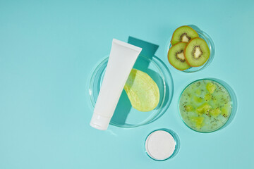 In the middle of the blue background photo is a round glass tray with an unlabeled white tube placed on top, next to it is a dish of crushed kiwi and sliced kiwi. From above view photo, copy space
