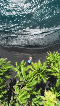 Vertical Screen: Vertical Aerial View of a Black Sand Beach with a Colorful Umbrella and Clear Ocean on the Big Island, Hawaii. The Scene Highlights the Beautiful Contrast Between Dark Sand 