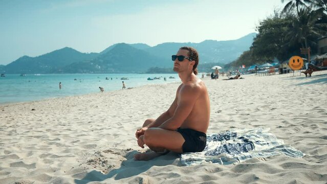 Millennial sitting sunbathing on beach and looking at seascape. Warm sand sound of surf and alight breeze from sea impress man with beauty. Calm man immersing himself process relaxation, enjoying sea.