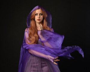 close up portrait of beautiful female model with long red hair, wearing a purple fairy  fantasy cloak with hood and gown. Elegant gestural hand poses as if casting magic spell. Isolated on dark black 