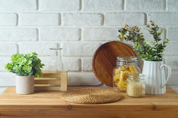 Empty wooden kitchen table with wicker place mat, food jars and plants over white brick wall ...