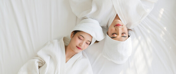 Two asian women are lying relax on a bed in white robes and towels wrapped around their heads. Top...