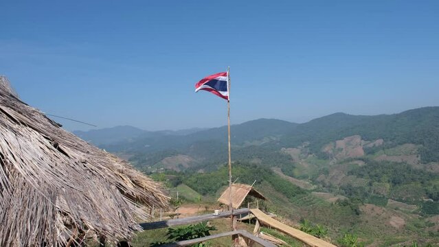 The Thai flag flutters in the wind. There is a complex mountain background.