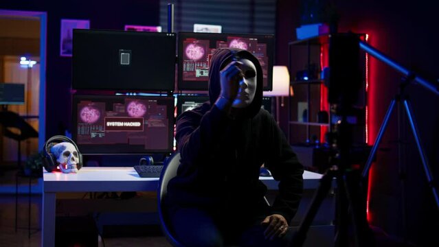 Hacker group member wearing anonymous mask filming video threatening another country government to steal military secrets, doing international espionage in hidden underground base, camera B