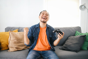 Excited Asian man playing video games using joystick while raising fist for winning the game at home