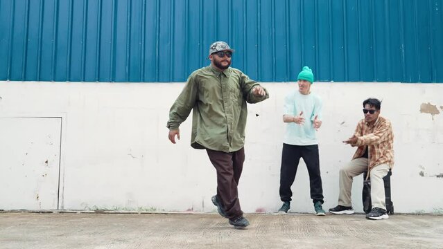 Skilled hipster doing cart wheel while multicultural friends encourage him. Hip-hop dancer moving foot step to music surrounded by group of hipster at wall near street. Outdoor sport 2024. hiphop.