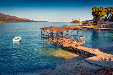 Sunny summer view of Ksamil resort with old wooden pier. Bright seascape of Ionian sea with white fishing boat. Calm outdoor scene of Albania, Europe. Vacation concept background.