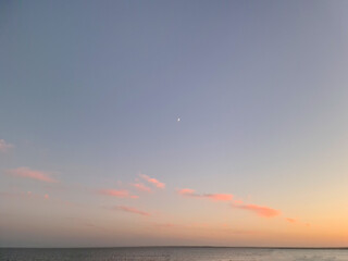 Obraz premium Little moon hangs in the sky over the ocean during dusk, accompanied by a vibrant afterglow. A beautiful natural landscape at sunset.