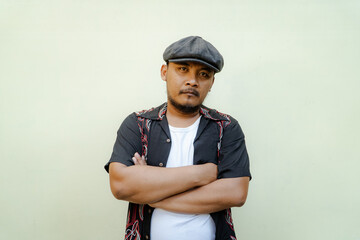 Macho man with beard and mustache looking serious isolated on beige background. Half body portrait of a vintage style Southeast Asian adult man in a patterned shirt, white tank top and newsboy cap