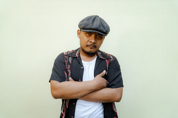 Macho man with beard and mustache looking serious isolated on beige background. Half body portrait of a vintage style Southeast Asian adult man in a patterned shirt, white tank top and newsboy cap