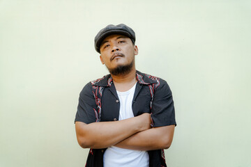 Macho man with beard and mustache looking serious isolated on beige background. Half body portrait of a vintage style Southeast Asian adult man in a patterned shirt, white tank top and newsboy cap