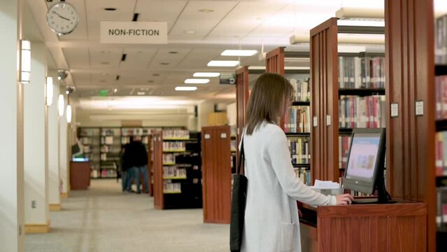 A woman with a bag uses a self-checkout kiosk in a library's non-fiction section, surrounded by bookshelves and a clock on the wall.