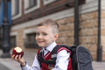 Little schoolboy sitting on a curb near school  with backpack, eating an apple as a snack after...