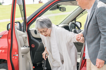 自動車の助手席から降りる杖をつく高齢者女性と介助する男性
