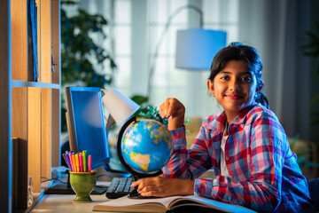 Asian Indian girl child studying at home on study table with computer, books, Globe model, victory...