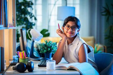 Asian Indian girl child studying at home on study table with computer, books, Globe model, victory...