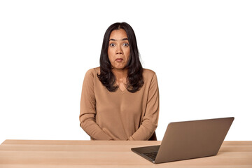 Filipino woman at desk with laptop shrugs shoulders and open eyes confused.