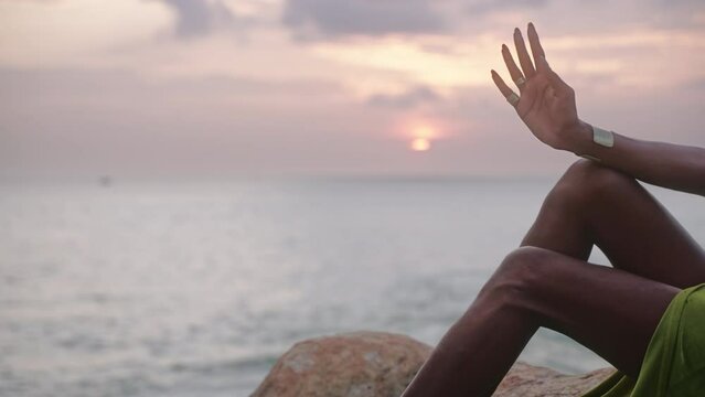 LGBTQ bipoc person hand with boutique golden jewelry poses against sun rays on cliff, ocean, looks aside. Divine gender fluid multiethnic model in seductive outfit stands on rock on sunrise.