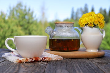 A white cup of hot tea next to a glass teapot and white jug with yellow flowers stands on a wooden...