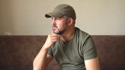 The man looks thoughtfully into the distance. Bearded face. Green baseball cap and clothes. Grey hair. In room. Reflections. Military. Close-up.