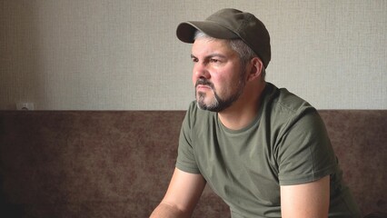 The man looks thoughtfully into the distance. Bearded face. Green baseball cap and clothes. Grey...
