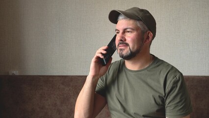 A man with a beard is calling on the phone. In hand there is a black smartphone. Green baseball cap...
