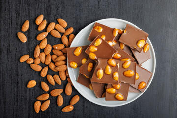 Broken chocolate bar with almonds on plate on black table. Top view.