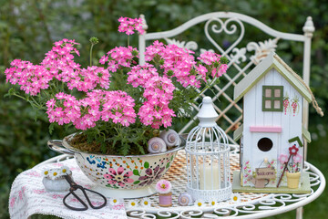 Garten-Arrangement mit pink Verbena im dekorativen Sieb, Vogelhaus und Kerze