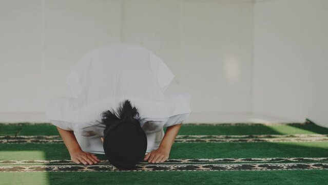 Asian muslim man in cap and thawb prostrating while praying to Allah on prayer mat at mosque