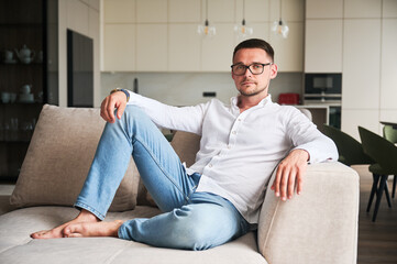 Charming man relaxing on couch in his apartment. Portrait of man in casual clothes and glasses lying on sofa on background of modern kitchen.