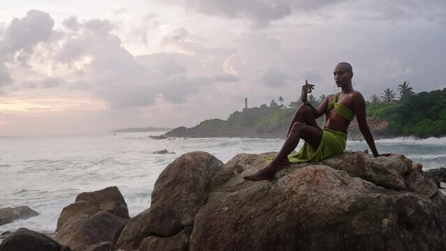 LGBTQIA black person in open dress, jewelry sits on rock on stunning ocean coastline sunset. Gender fluid ethnic divine model in seductive outfit fluttering in the wind against dramatic sky.